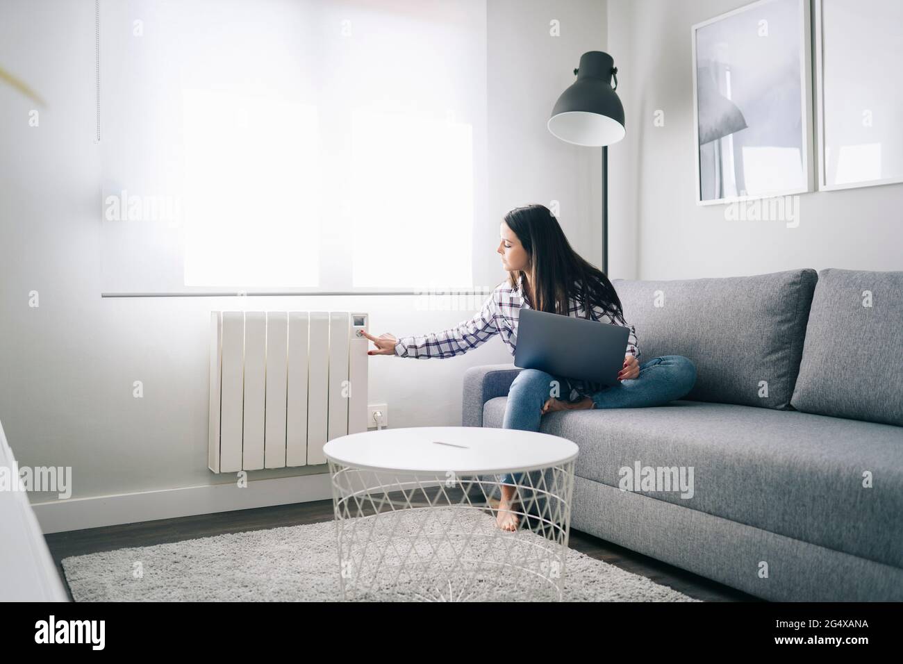 Donna che opera il riscaldamento mentre si siede con un computer portatile in soggiorno Foto Stock