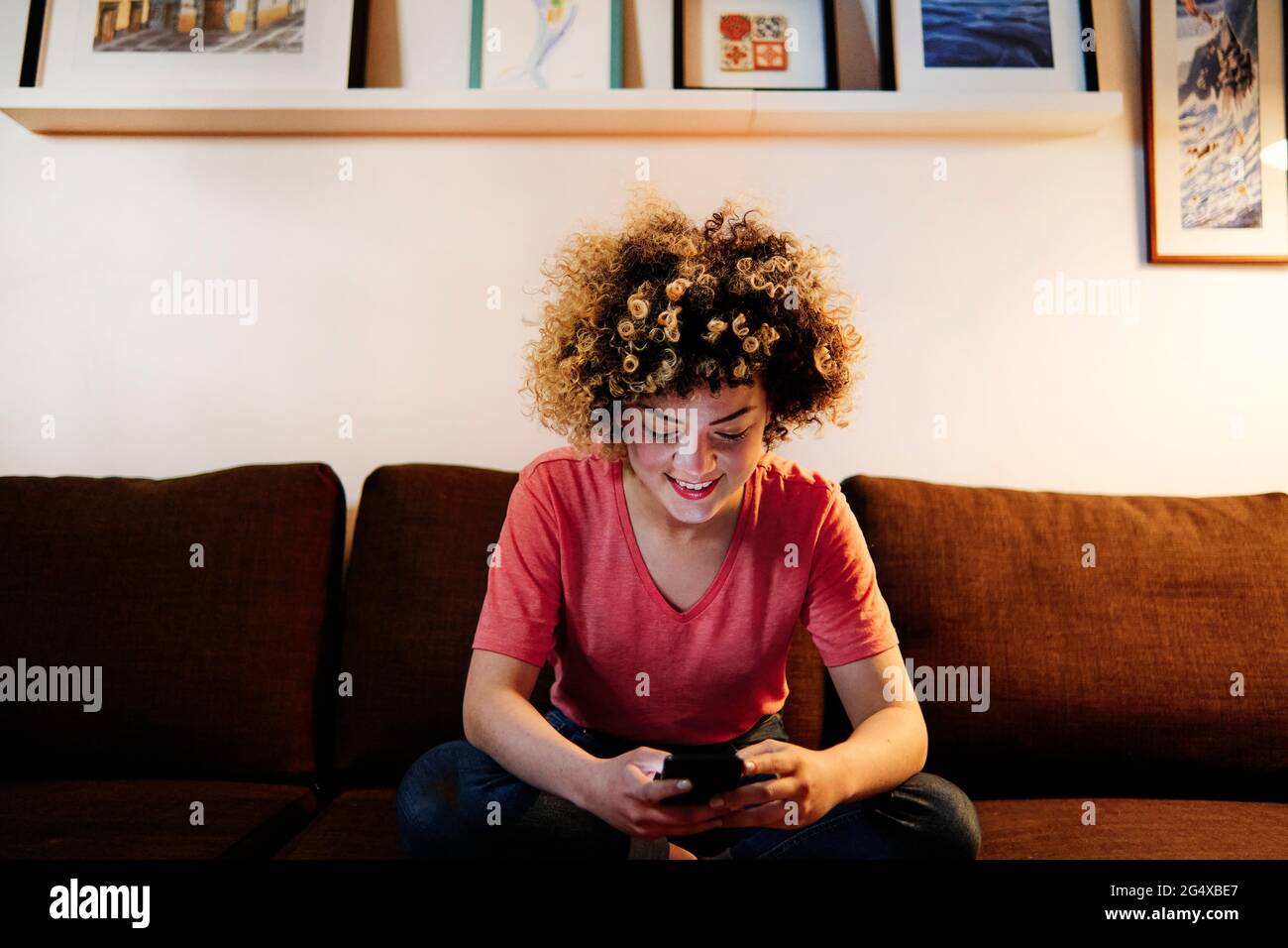 Sorridente giovane con messaggi di testo ricci tramite smartphone in soggiorno Foto Stock