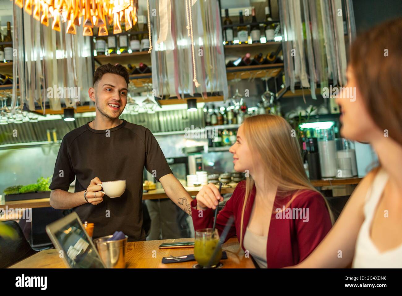 Un giovane sorridente in piedi con una tazza di caffè che parla con le amiche seduti al tavolo al bar Foto Stock