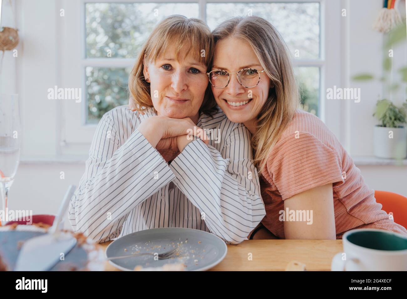 Figlia sorridente abbracciando la madre mentre si siede al tavolo da pranzo Foto Stock