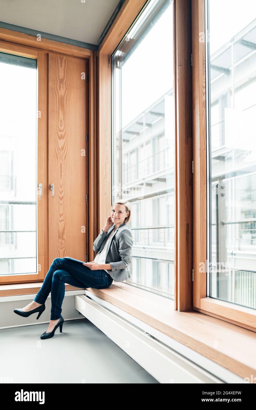 Donna d'affari sorridente seduta con gambe incrociate al ginocchio sulla soglia della finestra Foto Stock