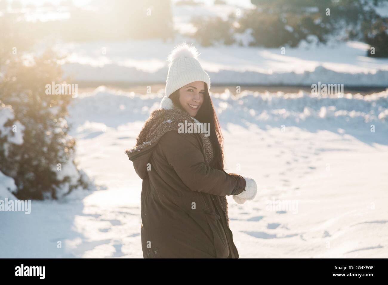 Donna sorridente che si affaccia sulle spalle mentre si trova sulla neve durante il giorno di sole Foto Stock