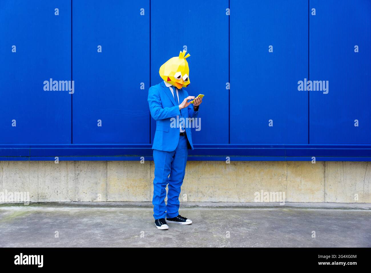 Uomo che indossa una vivace tuta blu e una maschera di protezione contro la parete blu con lo smartphone in mano Foto Stock