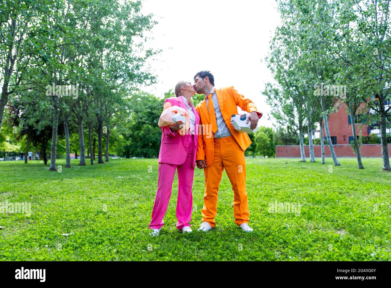 coupleÂ adulto indossa abiti vivaci che si baciano nel parco pubblico con maschere animali nelle mani Foto Stock