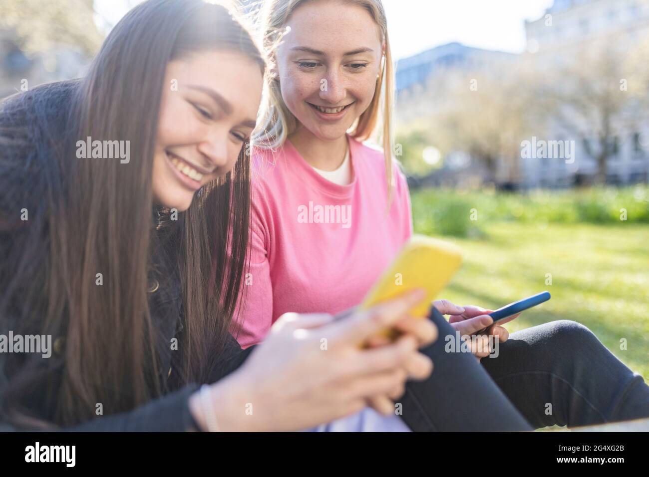Donna sorridente con capelli marroni messaggi di testo da amico femmina nel parco Foto Stock