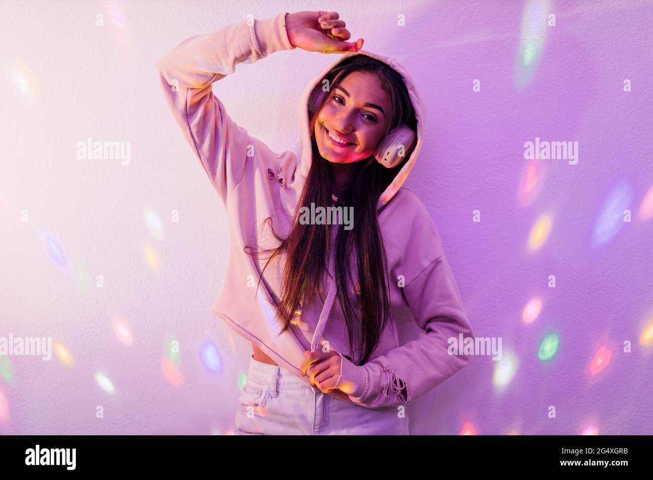 Bella cantante femminile che indossa la parte superiore con cappuccio mentre si ascolta la musica davanti alla parete in studio Foto Stock