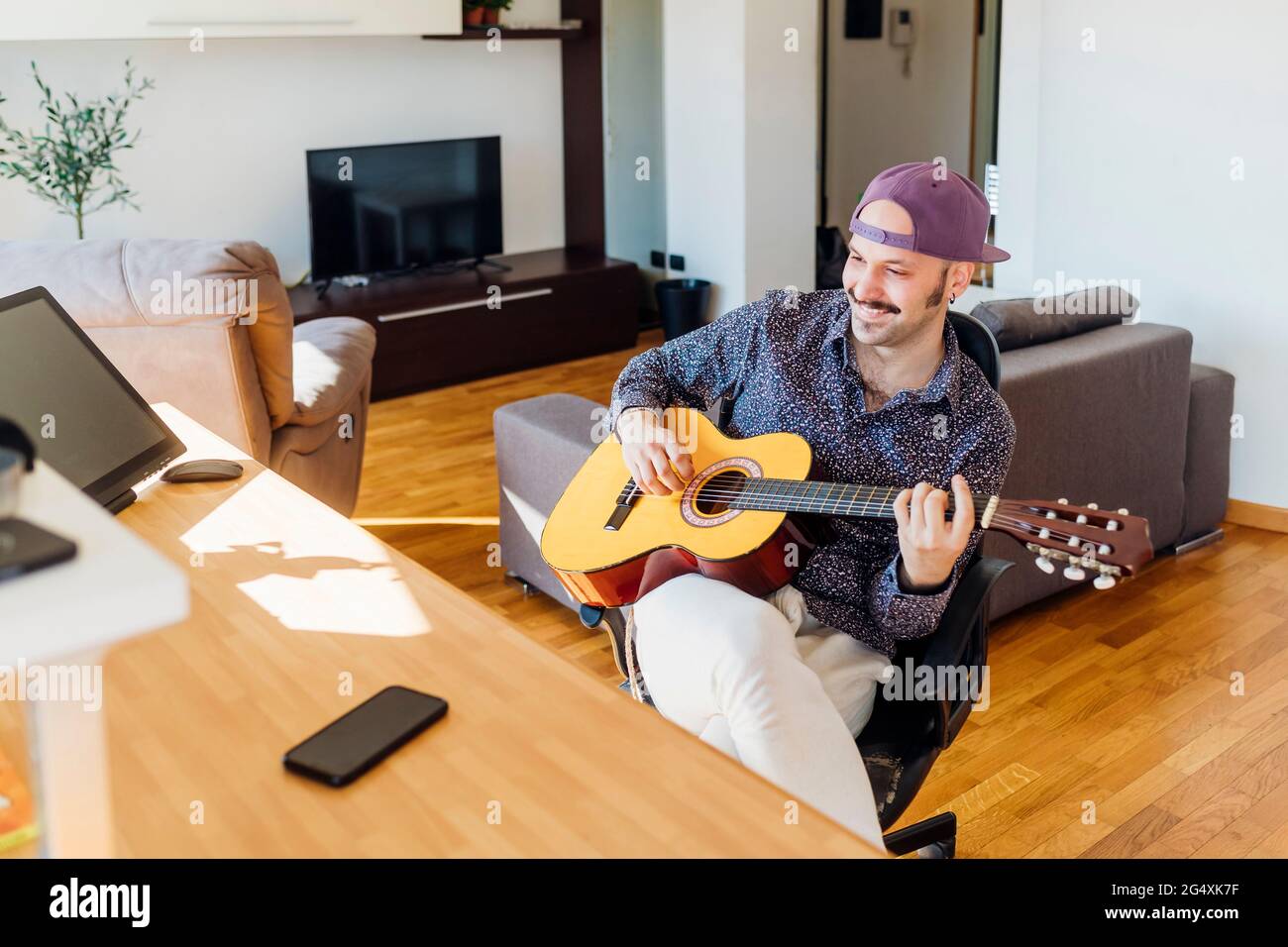 Un chitarrista maschio sorridente che indossa un cappello che suona la chitarra a casa Foto Stock