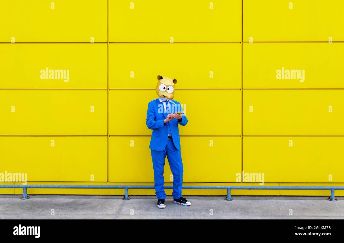Uomo che indossa una vivace tuta blu e una maschera di roditori davanti alla parete gialla con smartphone in mano Foto Stock