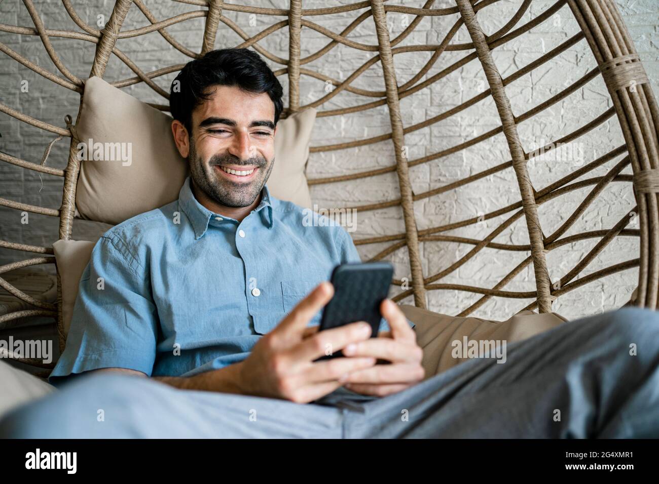 Uomo d'affari sorridente che usa il telefono cellulare mentre si siede sull'oscillazione di bambù Foto Stock