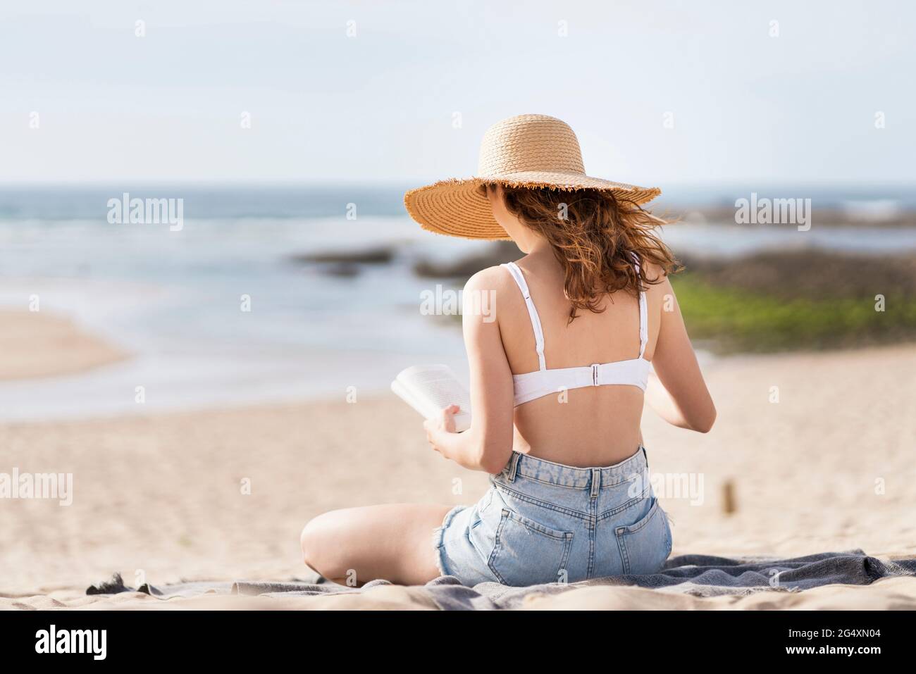 Donna che indossa cappello da sole leggendo libro mentre si siede in spiaggia durante le vacanze Foto Stock