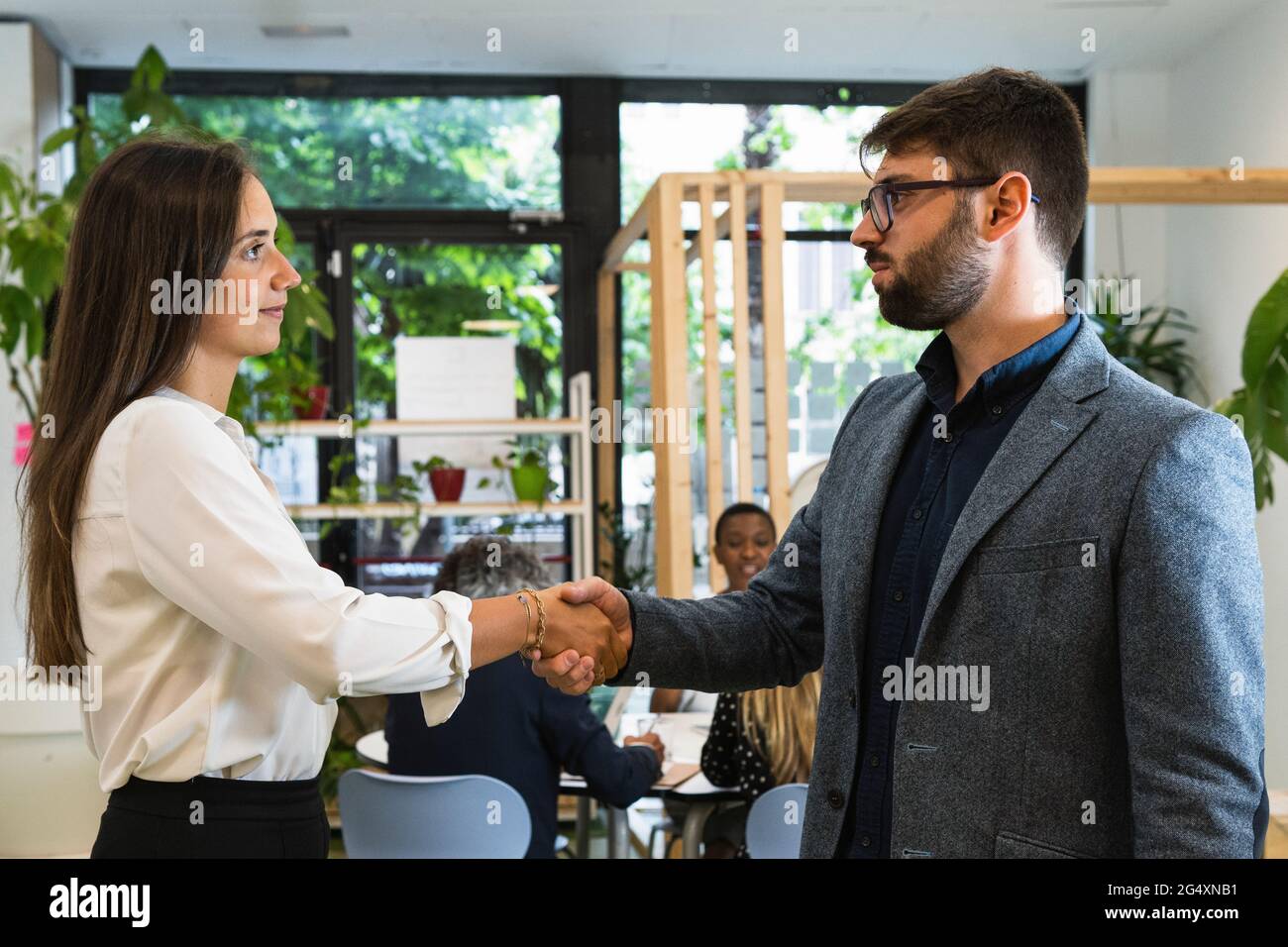 Giovani imprenditori maschili e femminili che fanno stretta di mano con i colleghi che discutono in background in ufficio Foto Stock