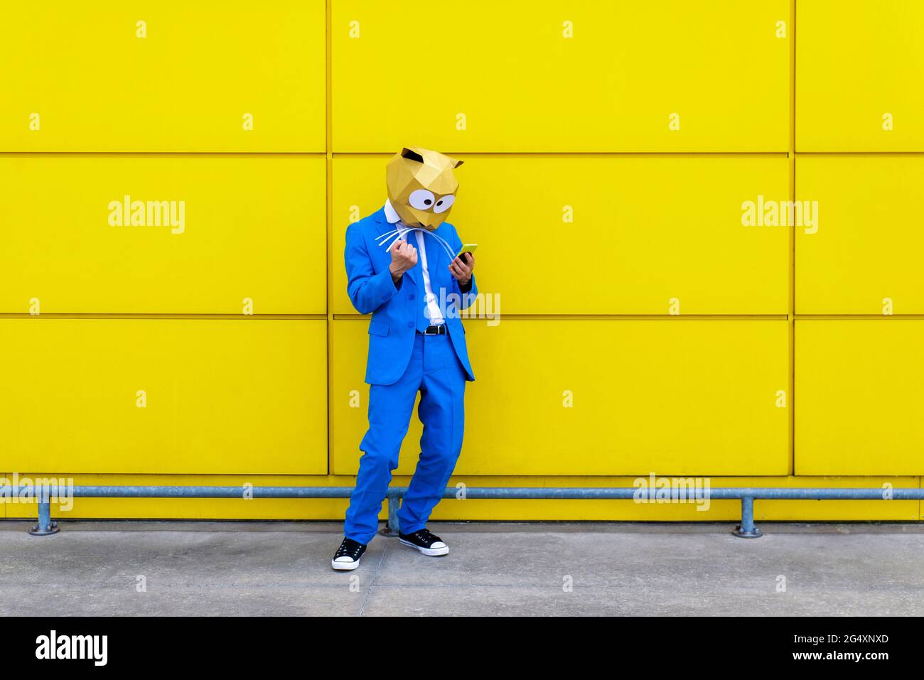 Uomo che indossa una vivace tuta blu e una maschera di roditori davanti alla parete gialla con uno smartphone in mano Foto Stock