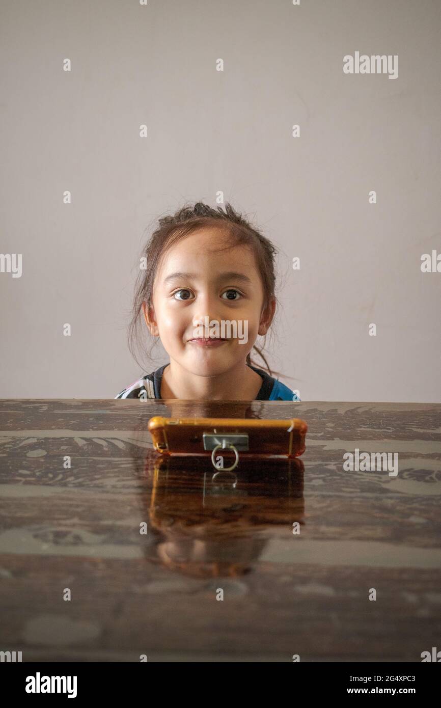 Ritratto di una bambina seduta al tavolo con il telefono cellulare davanti a lei. Guardare i video sul telefono. Foto Stock