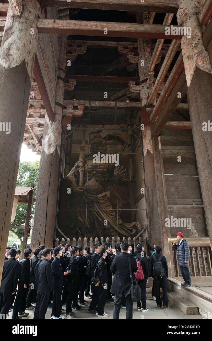 Gli scolari camminano nel tempio Todai passando davanti a una delle sue statue di guardiano a Nara, Giappone Foto Stock