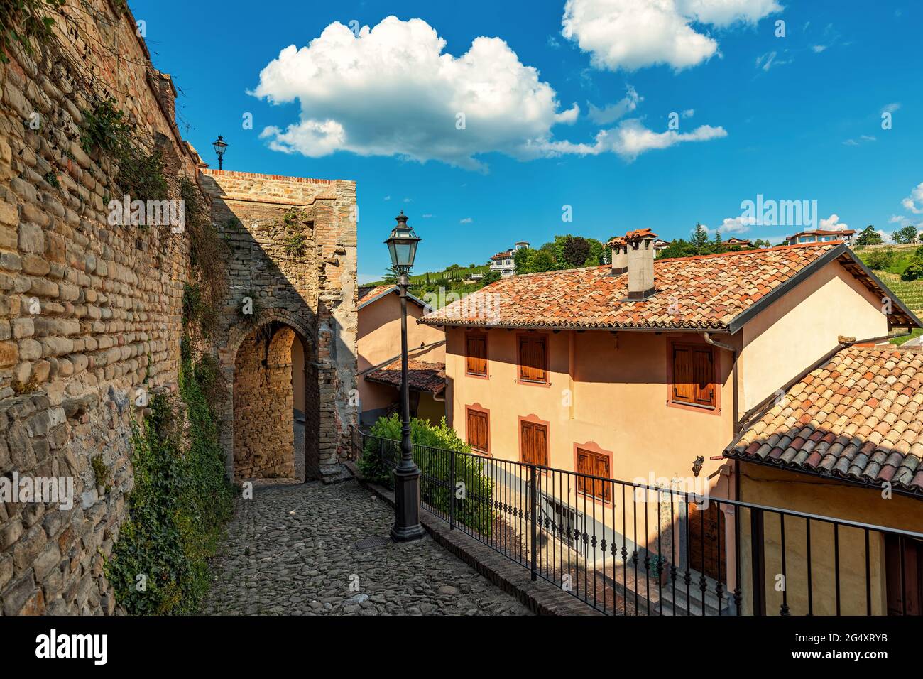Stretta strada acciottolata con lampione tra antiche mura circostanti e case sotto il bel cielo nella piccola città di Monforte d'Alba, Piemonte, Italia. Foto Stock