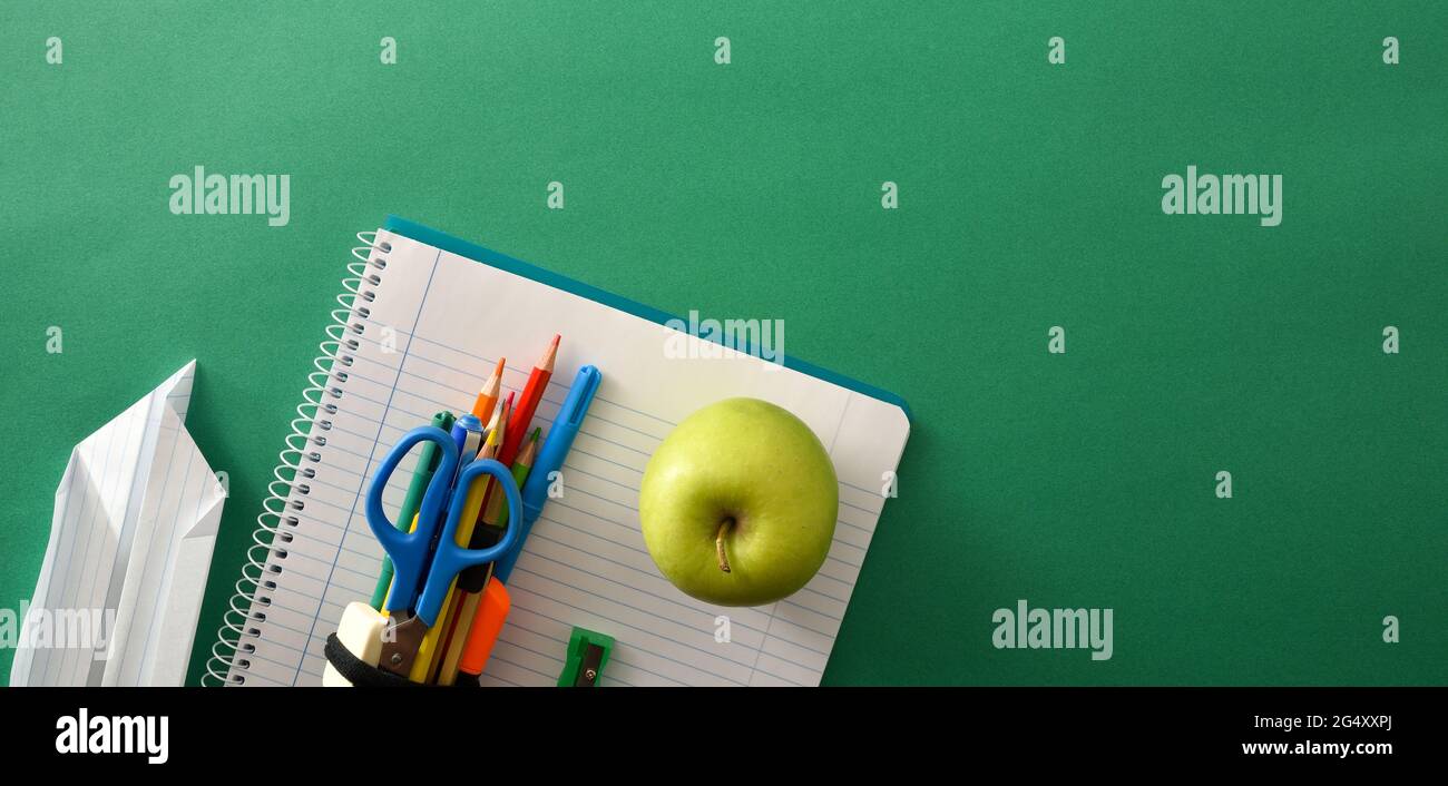 Strumenti per la scuola di notebook a spirale aperti sulla parte superiore con piano di carta e mela su sfondo verde con spazio per il testo. Vista dall'alto. Composizione panoramica. Foto Stock