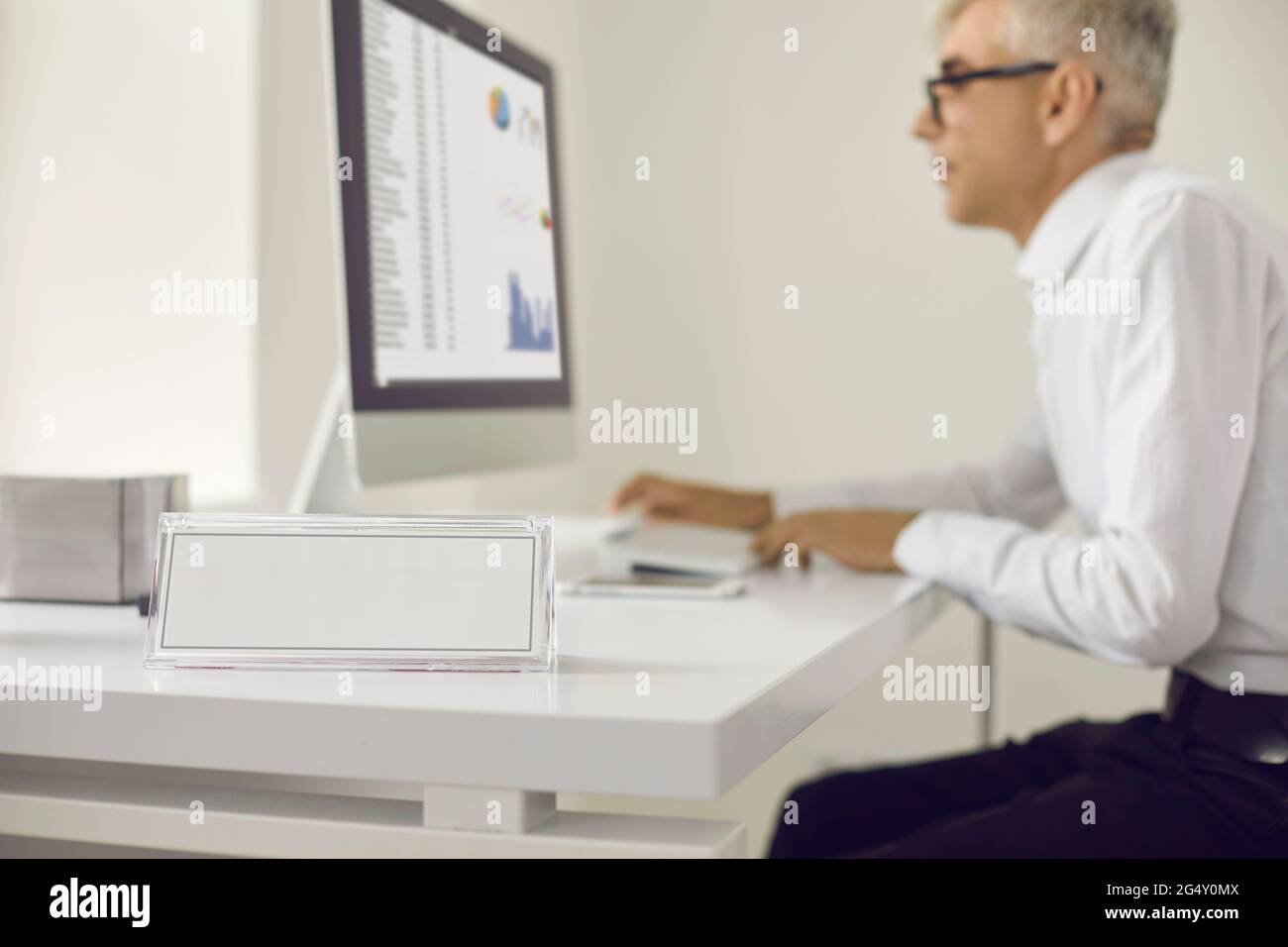 Primo piano della targhetta bianca vuota in ufficio sulla scrivania sullo sfondo di un uomo che lavora su un computer portatile. Foto Stock