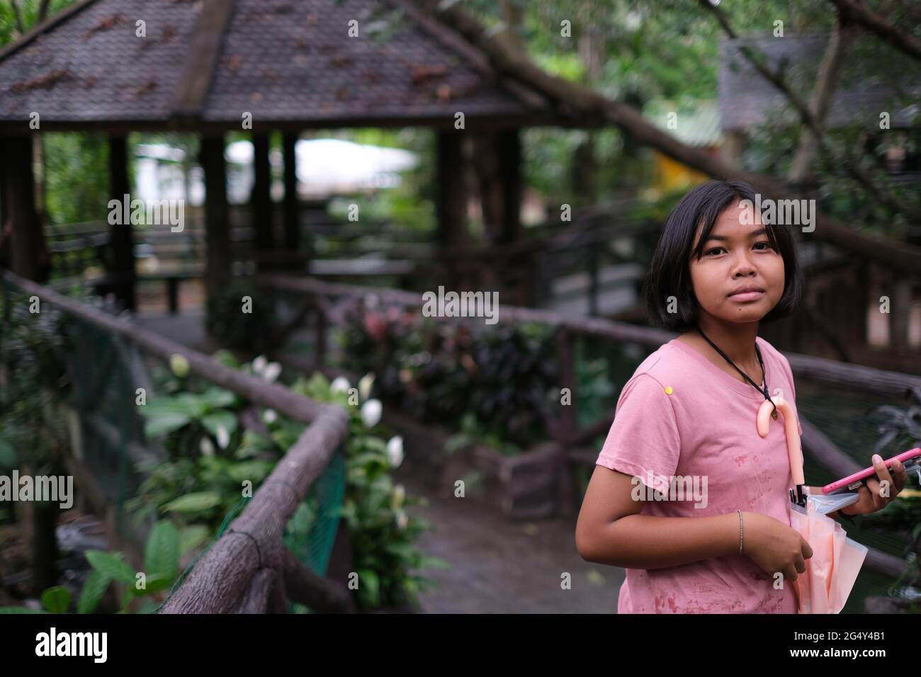 Una ragazza thailandese teen che guarda la macchina fotografica mentre cammina attraverso il giardino con case estive e sentiero in legno dopo la pioggia Foto Stock