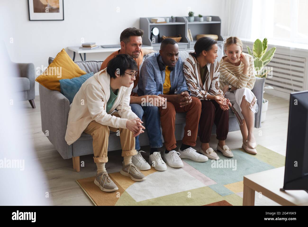 Vista completa su un gruppo multietnico di amici che guardano la TV a casa insieme mentre si siede in fila sul divano, spazio per le copie Foto Stock