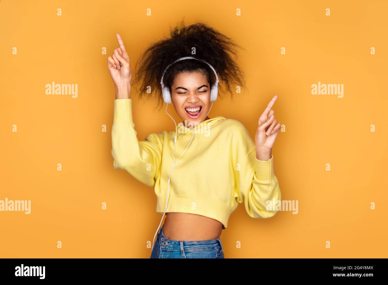 Gioiosa ragazza in cuffia, alza la mano in movimento danza e canta forte su sfondo giallo Foto Stock