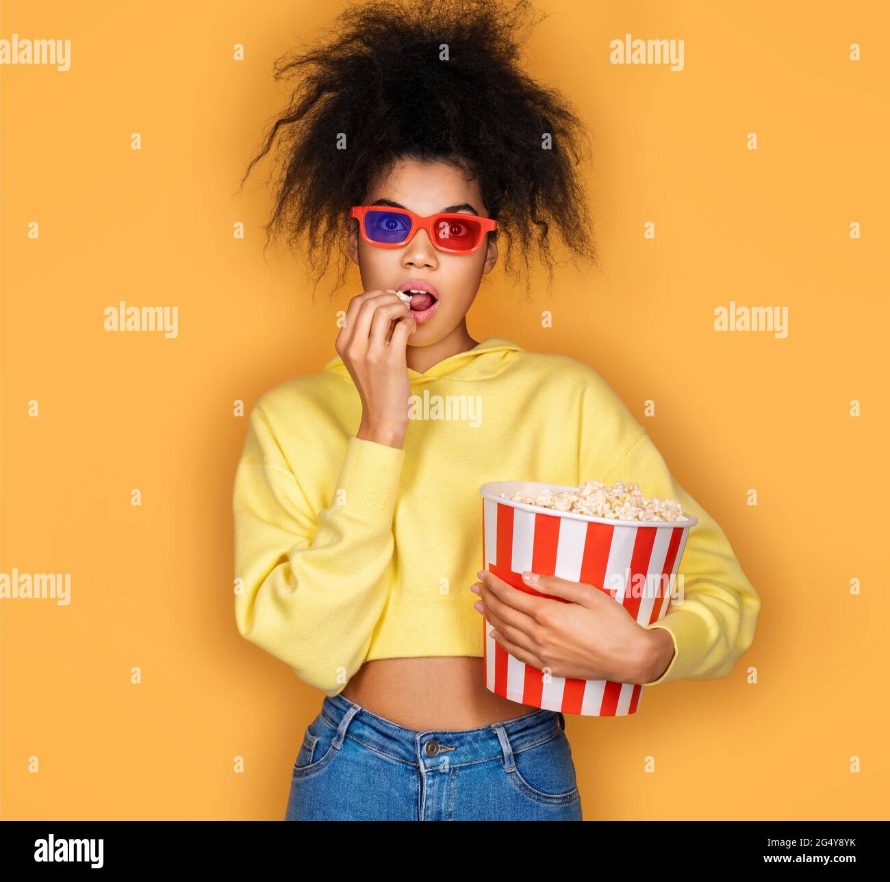 Giovane ragazza che guarda la tv con popcorn box. Foto di ragazza afroamericana su sfondo giallo Foto Stock