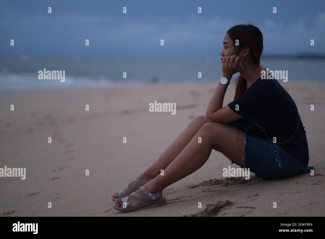 Giovane bella donna tailandese sta osservando il mare tempestoso su un tramonto, con il suo volto sepolto nelle mani Foto Stock