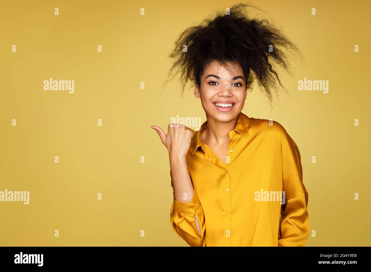 Sorridente ragazza punti da parte con il pollice, mostra sullo spazio di copia per il tuo testo. Foto di ragazza afroamericana su sfondo giallo Foto Stock