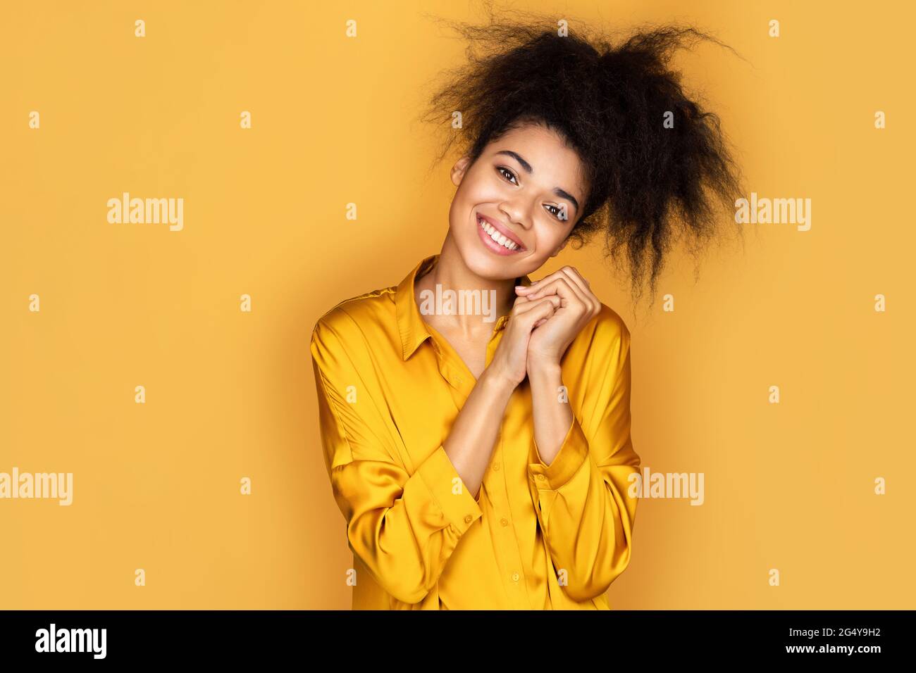 Bella ragazza sorridente tiene le mani pressate insieme sotto il mento, guarda qualcosa di carino. Foto di ragazza afroamericana su sfondo giallo Foto Stock