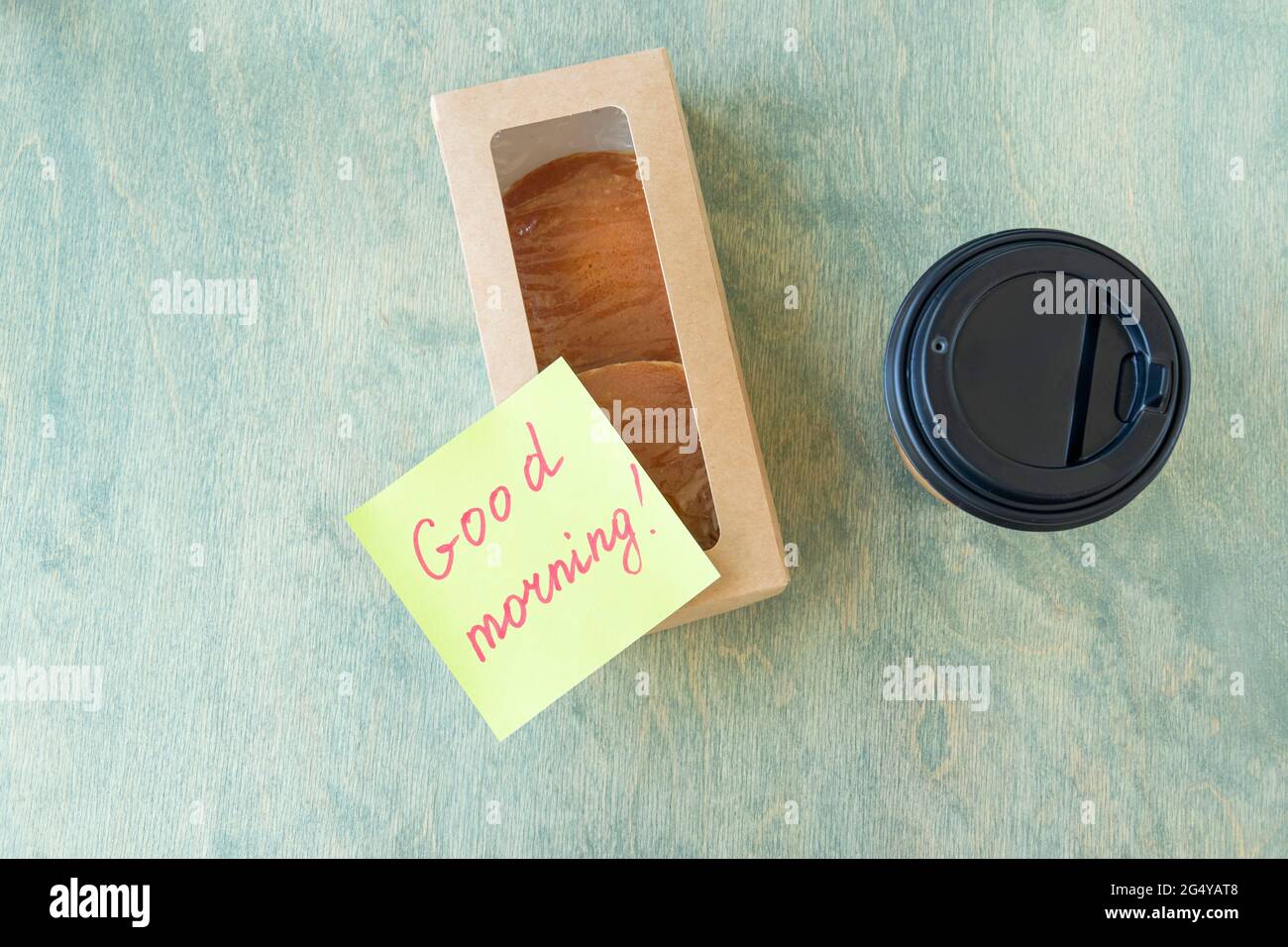 Vista dall'alto su una nota con le parole BUONGIORNO, frittelle in scatola di carta e caffè in tazza di carta su un tavolo di legno. Concetto di buon inizio di giornata. Disposizione piatta Foto Stock