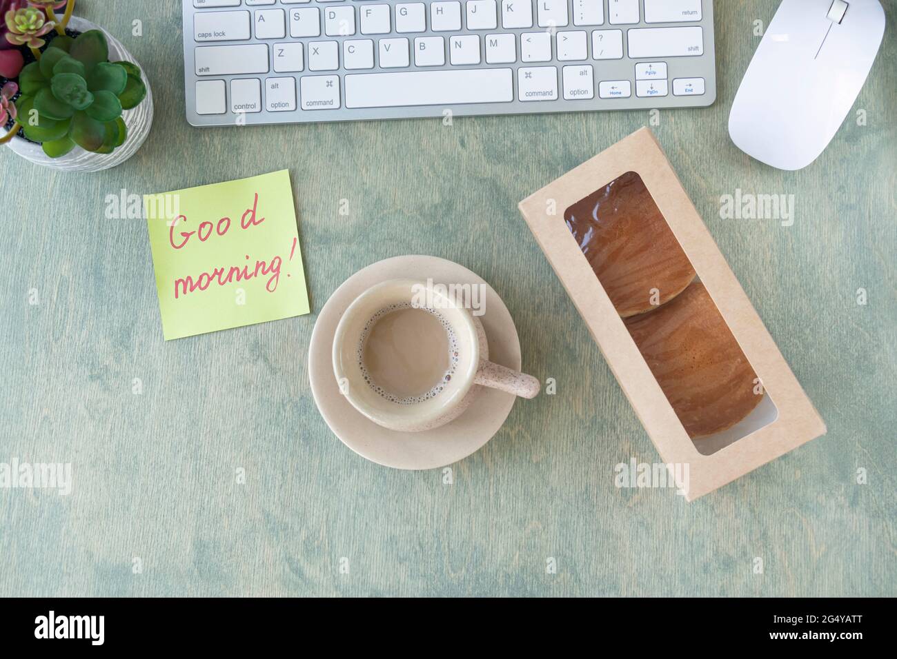 Vista dall'alto del desktop. Nota con le parole BUONGIORNO, frittelle in scatola di carta, tazza di caffè, tastiera, cactus e mause su un tavolo di legno. Concetto di buon inizio di giornata. Disposizione piatta Foto Stock