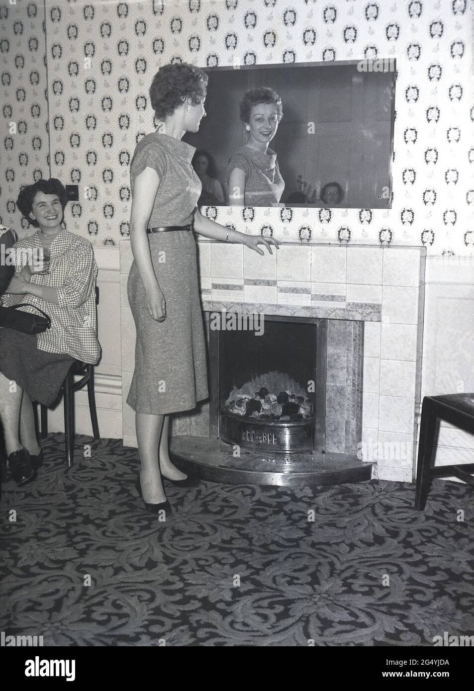 1957, storica, in una festa al Victoria Hotel, Leeds, una giovane donna che indossa un abito elegante in piedi accanto al camino che si affaccia su uno specchio. Foto Stock