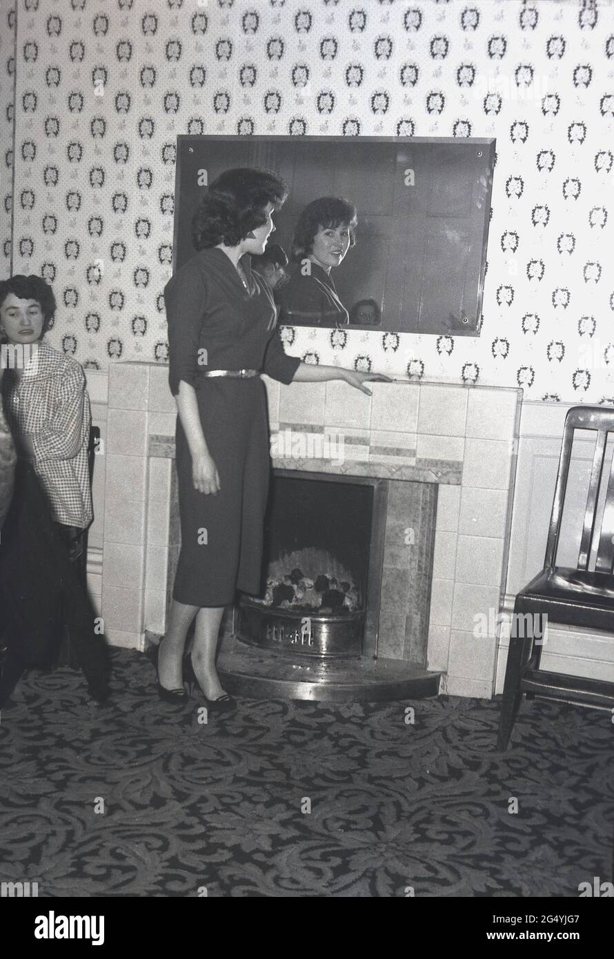 1957, storica, in una festa al Victoria Hotel, Leeds, una giovane donna che indossa un abito elegante in piedi accanto al camino che si affaccia su uno specchio. Foto Stock