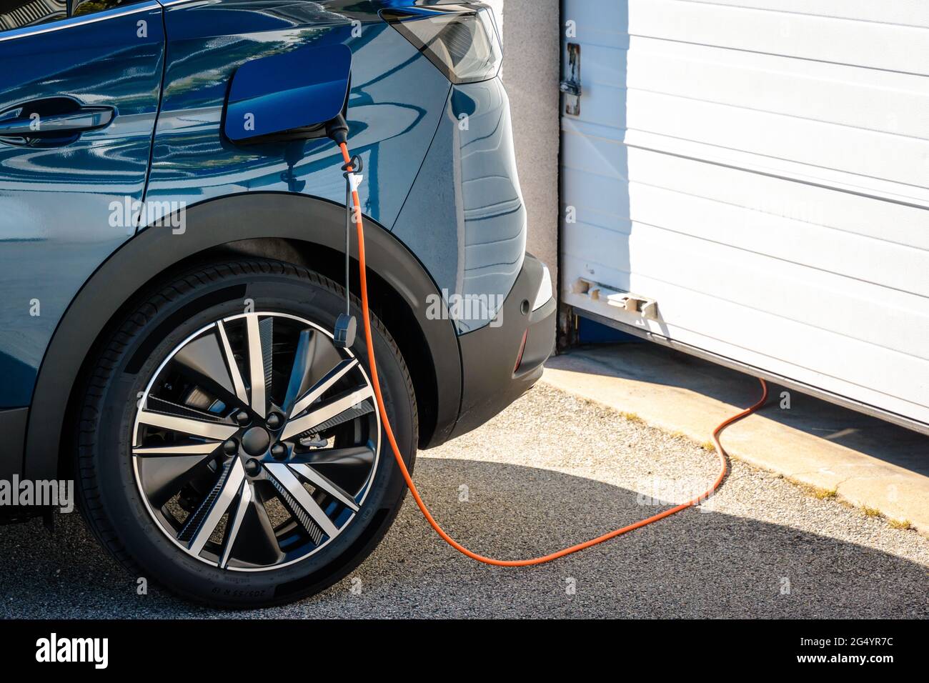 Un'auto elettrica ibrida parcheggiata davanti alla porta del garage è collegata tramite un cavo di ricarica per ricaricare la batteria. Foto Stock
