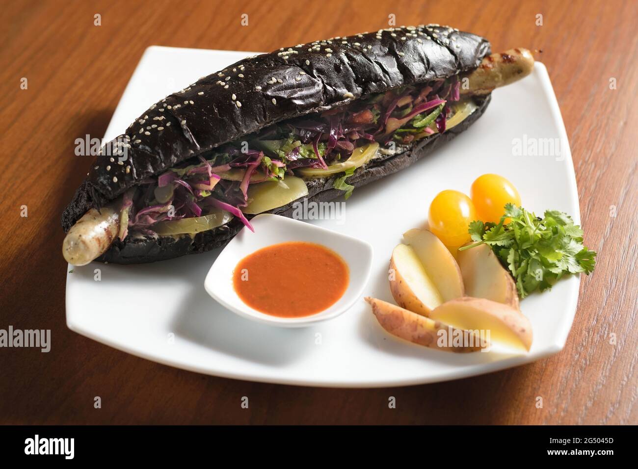 Hot dog nero lungo con salsiccia di pollo fritta con insalata, su un piatto con salsa, pomodori e patate Foto Stock