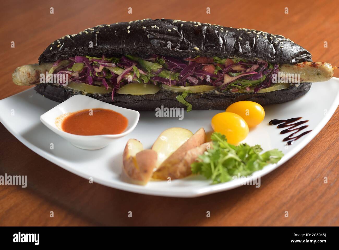 Hot dog nero lungo con salsiccia di pollo fritta con insalata, su un piatto con salsa, pomodori e patate Foto Stock