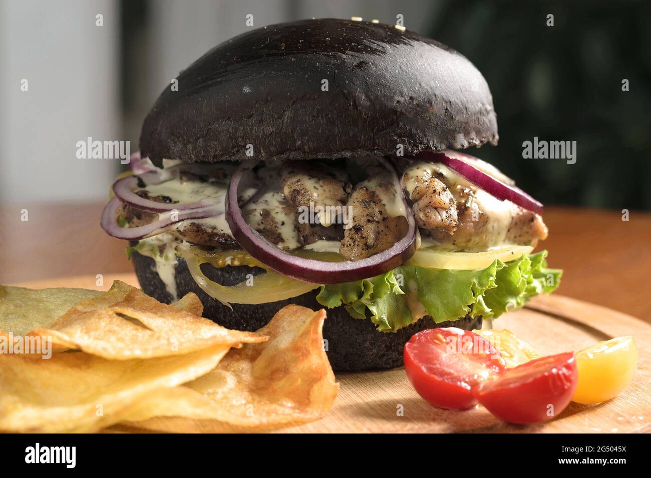 Grande hamburger nero primo piano con carne alla griglia, formaggio, anelli di cipolla e foglie di lattuga su tavola di legno, con patatine, con ciliegia gialla e rossa Foto Stock