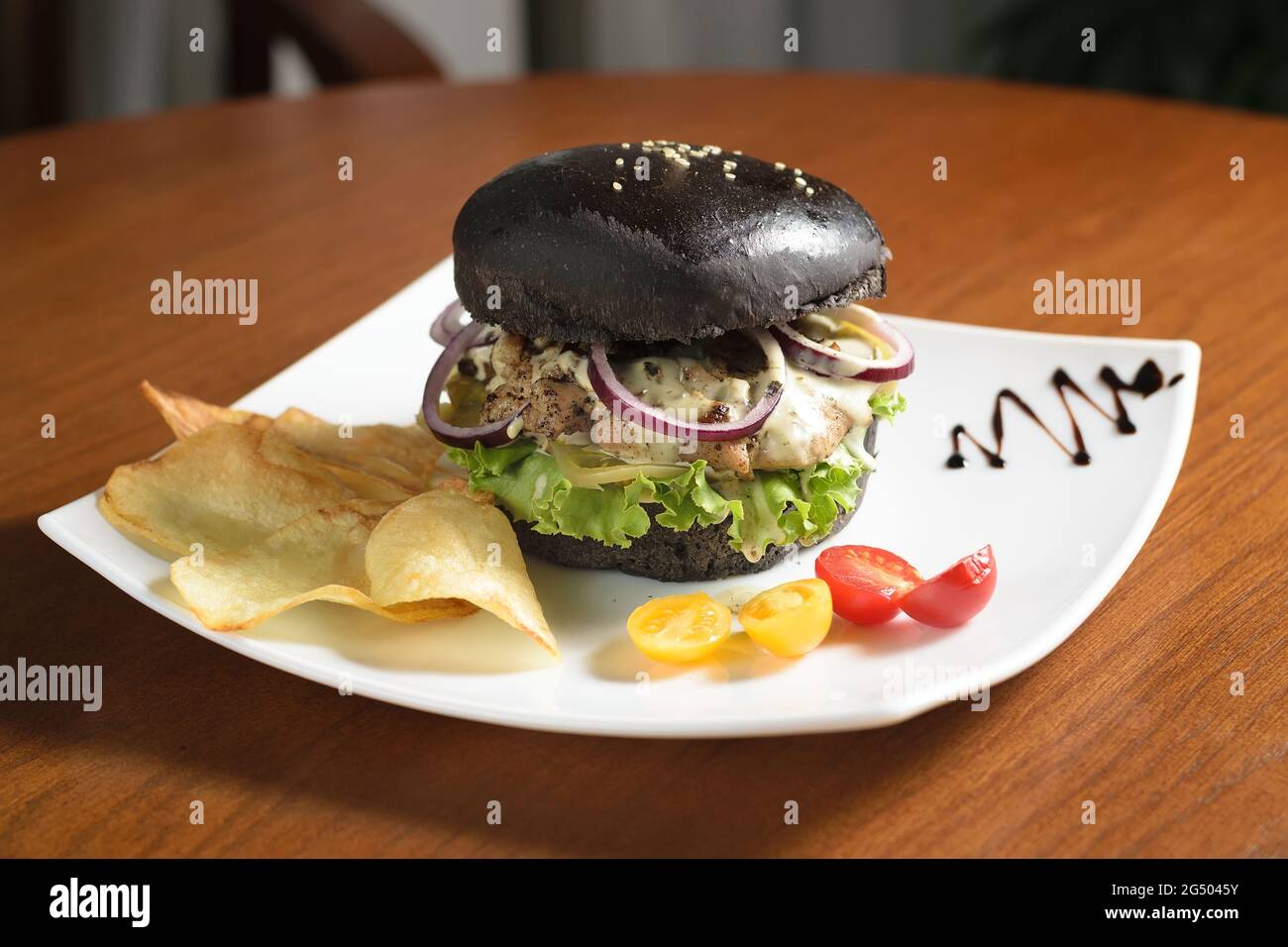 Hamburger nero grande con carne alla griglia, formaggio, anelli di cipolla e foglie di lattuga su un piatto bianco, con patatine, con pomodori ciliegini gialli e rossi Foto Stock