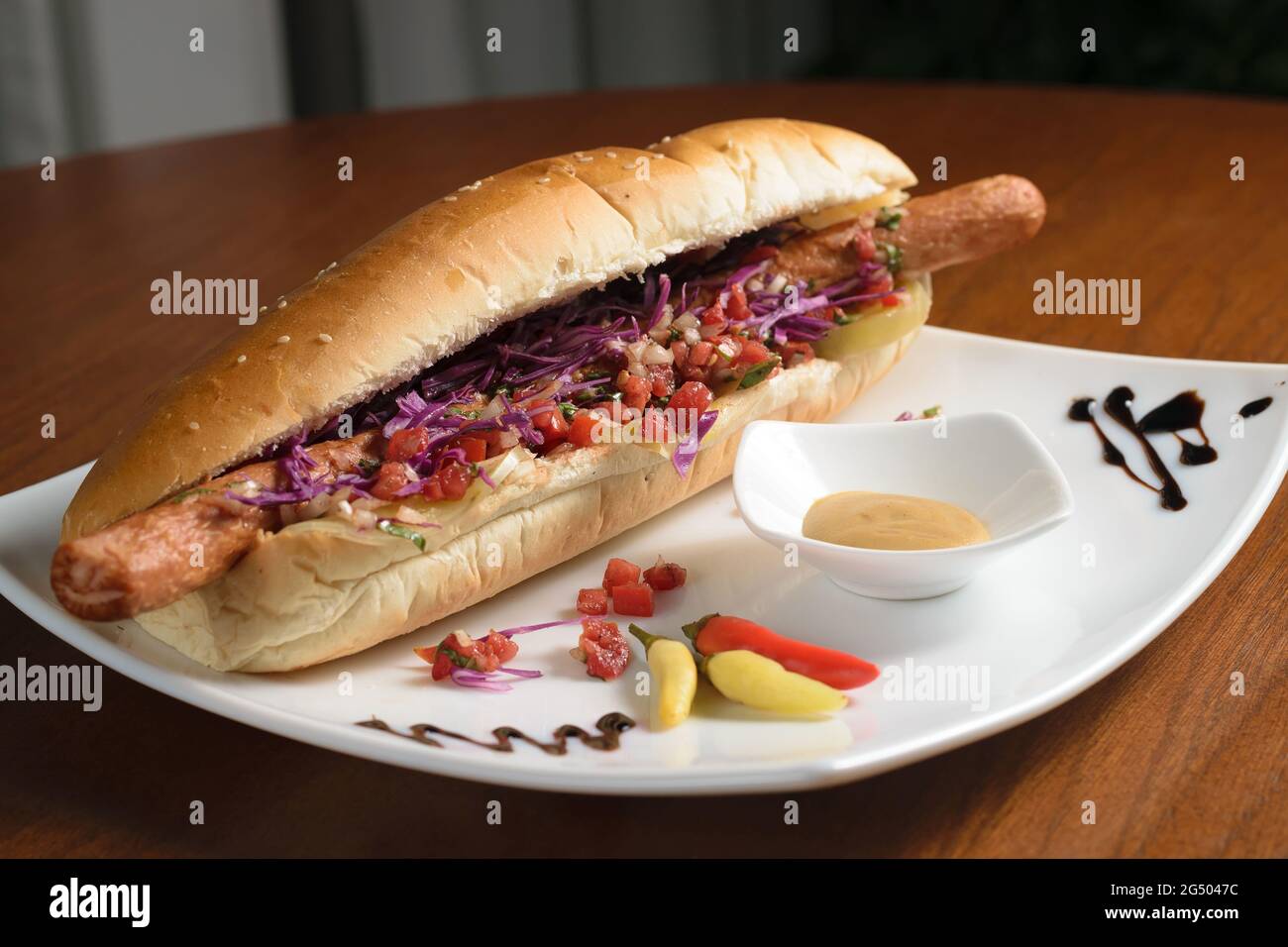 Hot dog lungo con due salsicce, cavolo e pomodori, e con fette di pomodoro verde salato, su un piatto bianco con senape e peperoncino Foto Stock