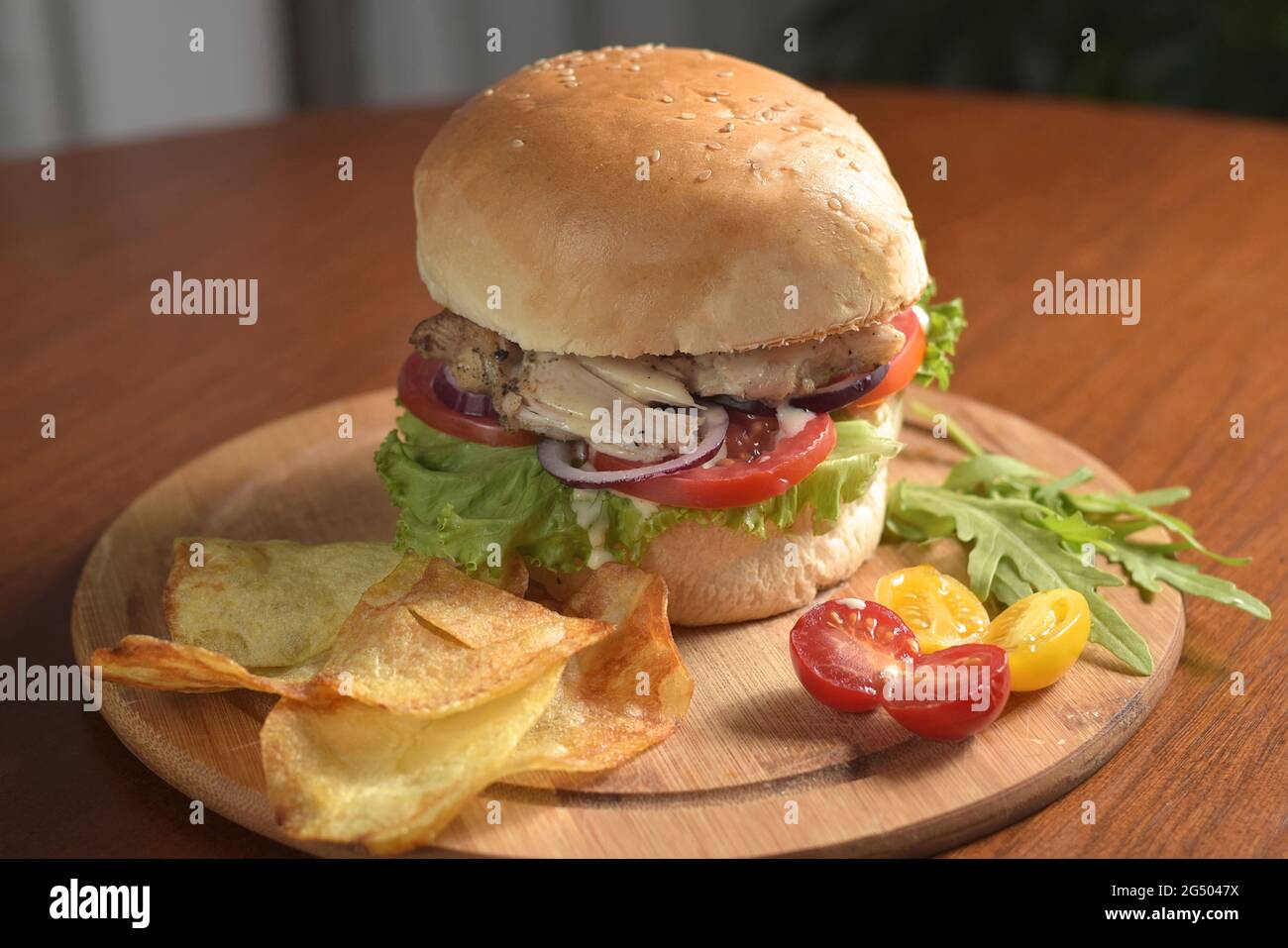 Grande hamburger primo piano con carne di pollo fritta, pomodoro, formaggio, anelli di cipolla e foglie di lattuga su una tavola di legno, con patatine, con giallo e rosso Foto Stock