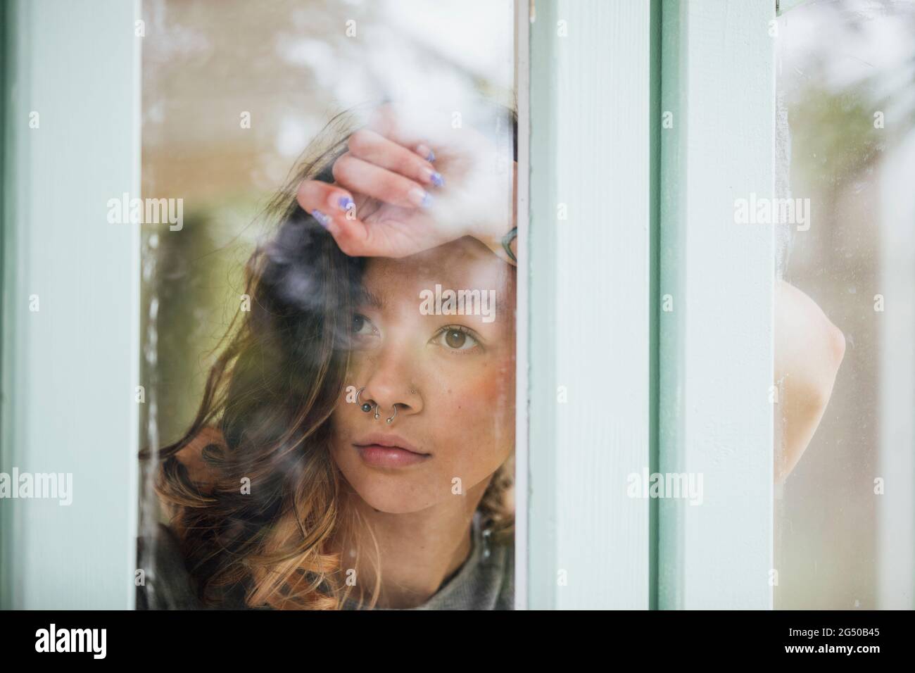 Una giovane donna, fotografata attraverso la finestra. Sta poggiando sul braccio sollevato contro la finestra. Foto Stock