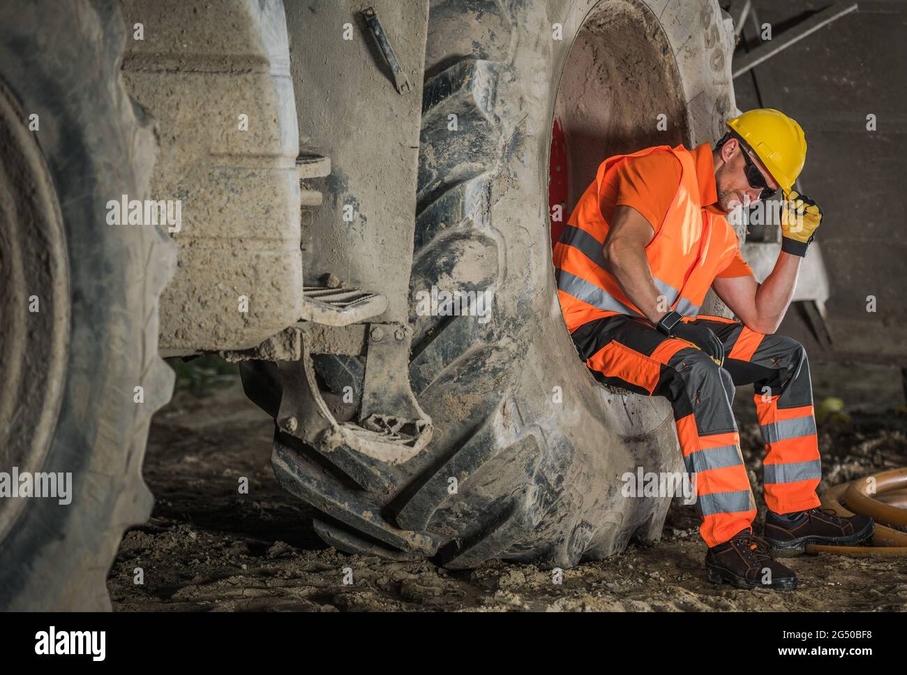 Un operaio caucasico della costruzione stradale nei suoi 40 anni che si trova a breve e a sedere all'interno di una ruota motrice per impieghi pesanti di grandi dimensioni. Area con cappello duro. Tema industriale. Foto Stock