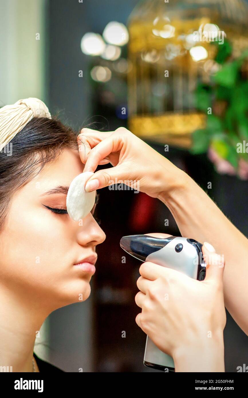 Vista laterale di un make-up artista utilizzando l'aerografo facendo un airbrush trucco fondazione su una faccia femminile in un salone di bellezza Foto Stock
