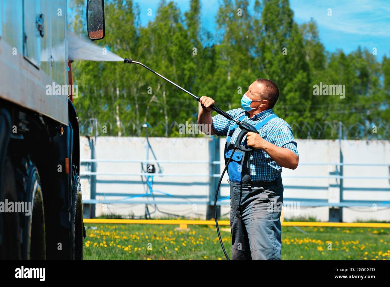 Autolavaggio per veicoli pesanti e cargo. Un uomo UN operaio del lavaggio dell'automobile in una maschera medica lava un camion con una pistola dell'acqua sulla strada in un giorno d'estate. Foto Stock