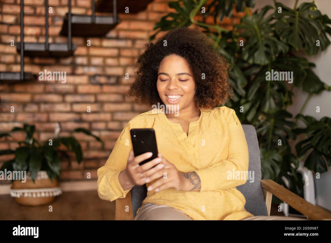 Gioiosa giovane afroamericana che usa uno smartphone seduto sulla poltrona di casa, sorridente donna nera ama chattare online, passare il tempo in social network Foto Stock