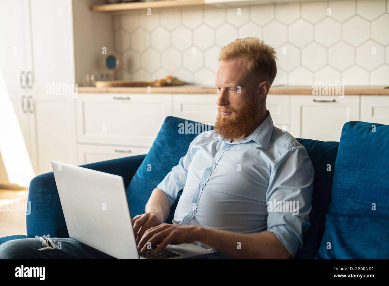 Uomo rosso focalizzato utilizzando il computer portatile per studiare o lavorare online, ragazzo freelance digitando seduto sul divano a casa, sviluppa nuovo progetto lavorando a distanza Foto Stock