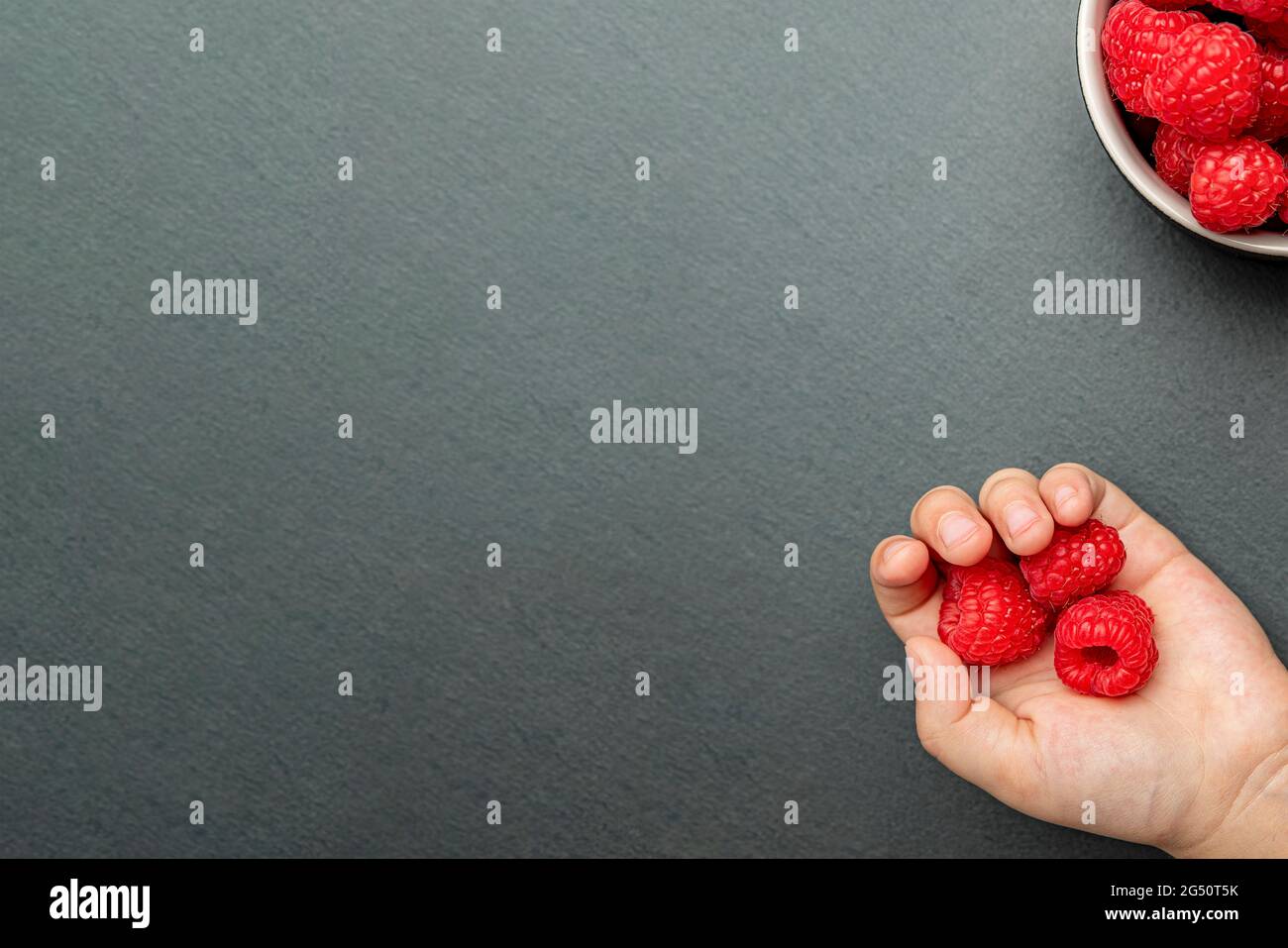 Il bambino prende un lampone e lo mette in mano. Bacche su sfondo nero in un piatto, posizionare per inserire testo, ricetta o disegno Foto Stock