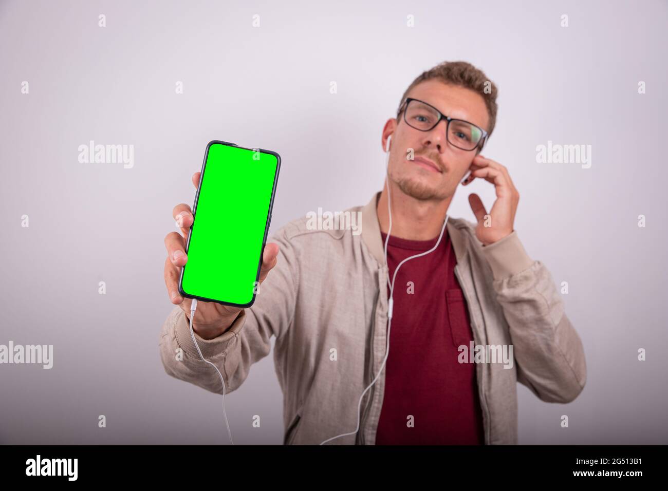 Un giovane e attraente uomo ascolta la musica con gli auricolari, mostrando lo schermo dello smartphone, lo schermo verde per inserire il suo logo, la foto dello studio. Foto Stock