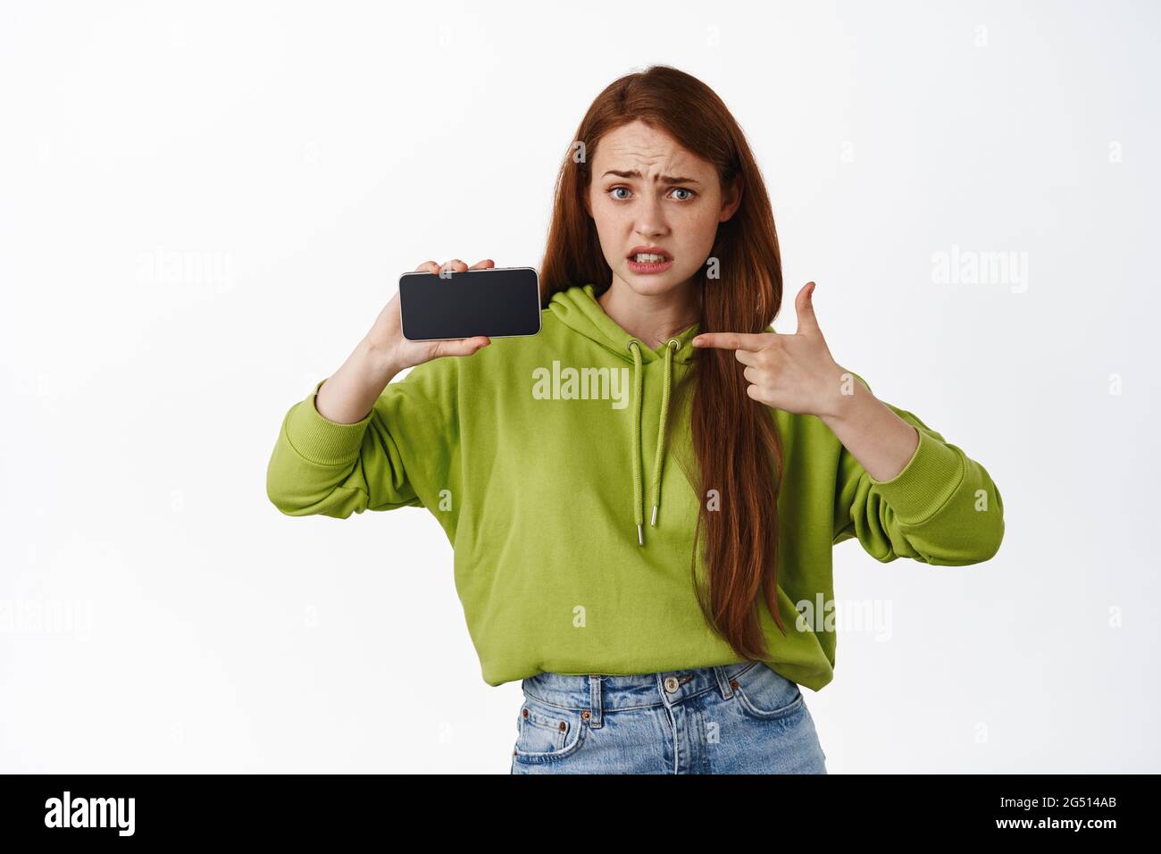Ritratto di girl punta allo schermo del telefono con la faccia interessata e delusa, avete visto questo, mostrando cattive notizie sul telefono cellulare, sfondo bianco Foto Stock