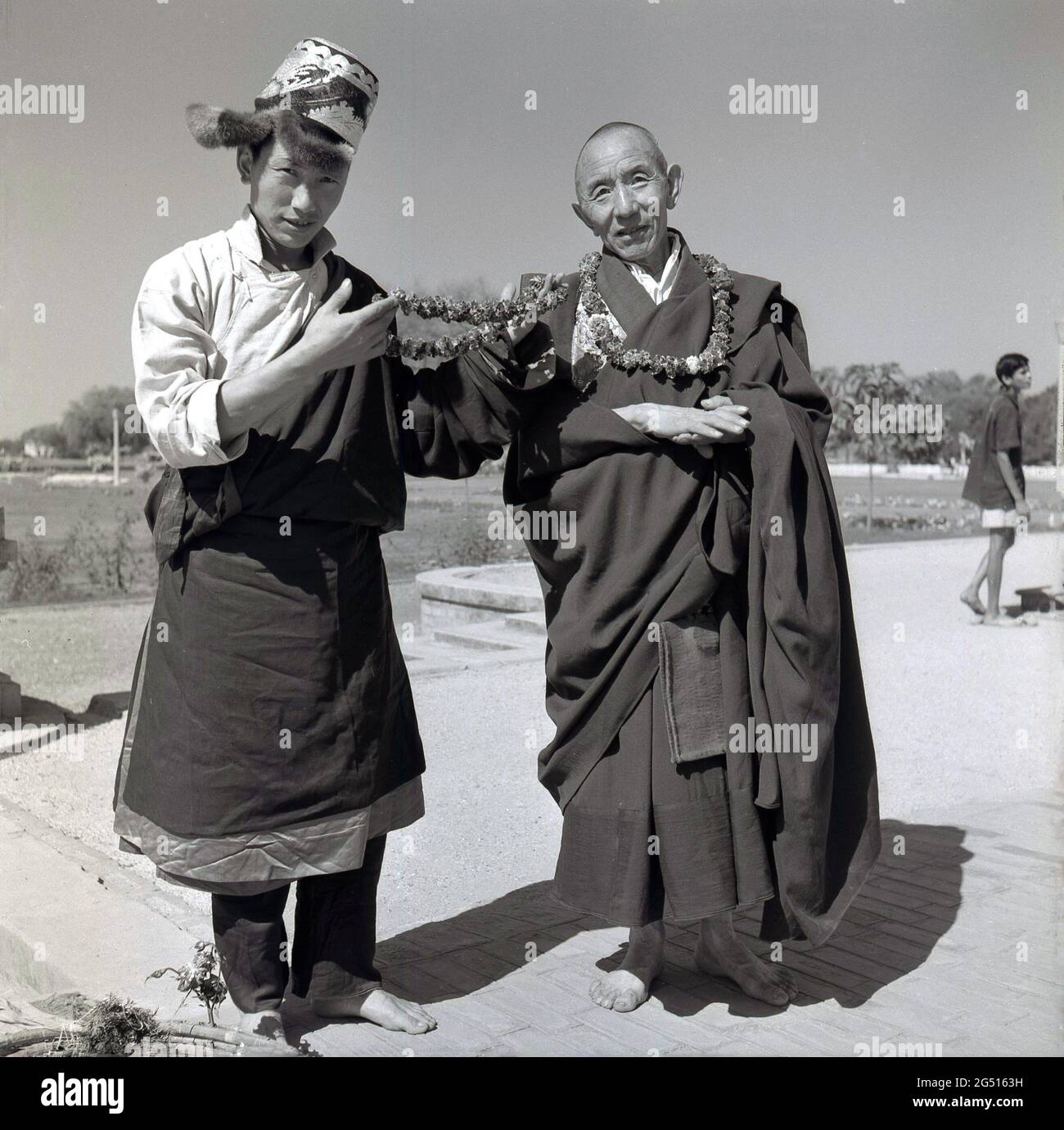 Anni '50, foto storica di J Allan Cash di un giovane in abiti e cappello, in piedi con un monaco fuori del tempio buddista, mulagandhakuti vihara, Sarnath, Varanasi, india. Il giovane tiene ghirlande o trecce di fiori, che sono indossati intorno al collo e che sta offrendo ai visitatori. Il tempio è stato costruito nel 1931 - sul sito dove il Buddha Gautama mediato durante la sua prima stagione piovosa - da Angarika Dharmapala ed è mantenuto e gestito dalla società Mahabodhi di cui ha fondato. Foto Stock