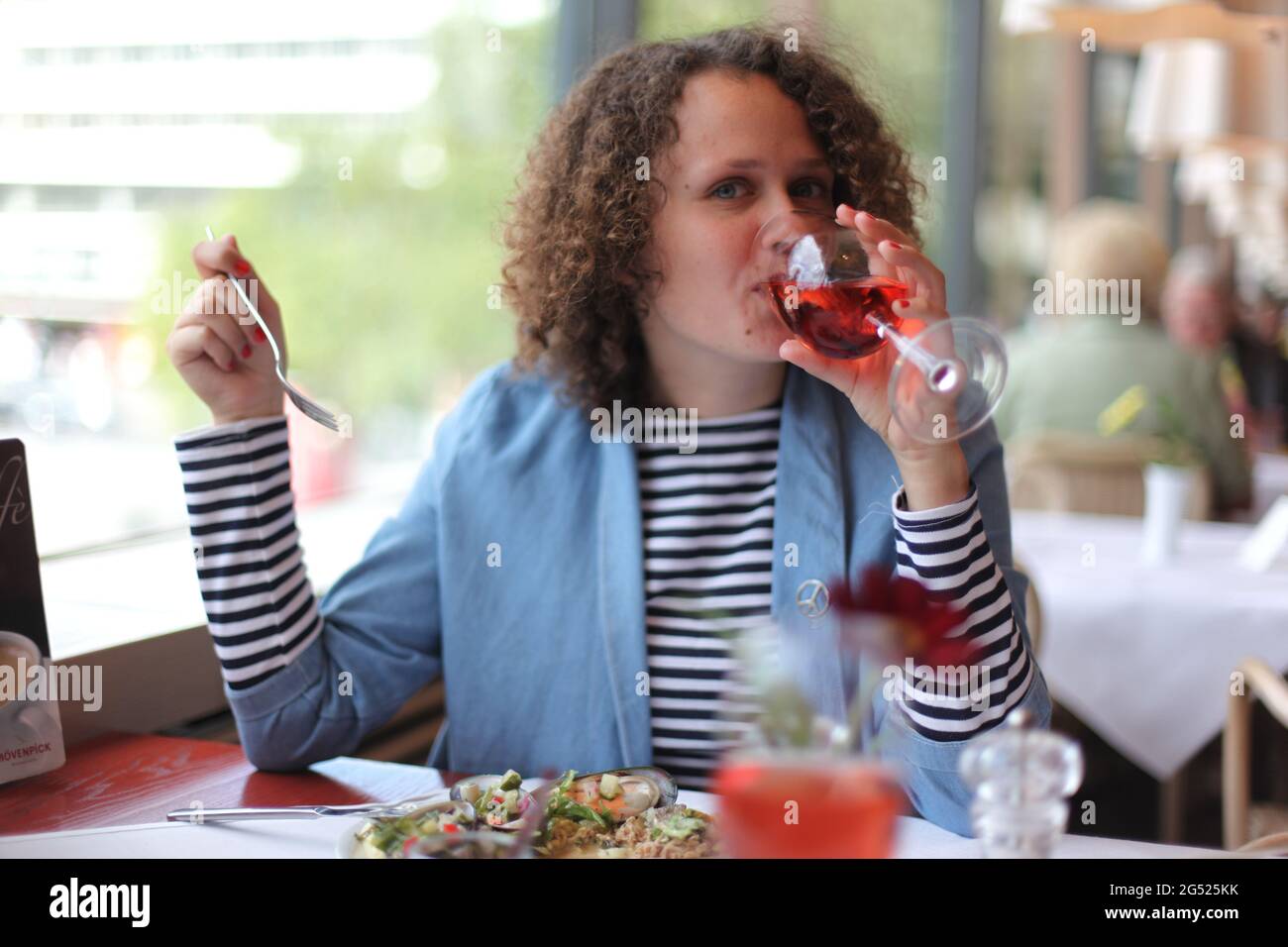 Felice giovane donna di stile in abito informale bere vino e guardando la macchina fotografica mentre si siede al tavolo durante il pranzo al ristorante Foto Stock